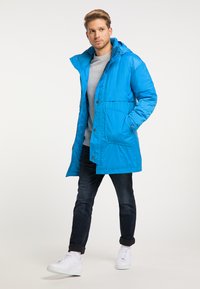 Blå polstret jakke med hætte, med trykknapper og forlommer, båret over en grå sweatshirt og parret med mørke jeans og hvide sneakers.