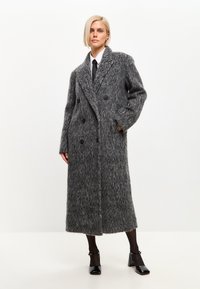 Abrigo gris de doble botonadura con un material texturizado y suave. Largo, con solapas de muesca y dos botones negros. Llevado sobre una camisa blanca y una corbata.