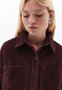 Burgundy kordflorskjorta med krage, två bröstfickor och bruna knappar; har en vertikal ribbad struktur.