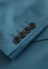 Quatre boutons noirs cousus sur le poignet d’une manche de blazer bleu sarcelle texturé, avec des boutonnières assorties visibles.