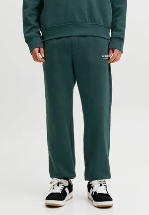 Homme portant un pantalon de survêtement vert foncé et un sweat assorti avec des baskets noires et blanches, debout devant un fond uni.