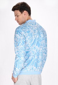 Pulover bleu deschis cu model botanic alb, guler și manșete cu nervuri. Țesătură netedă cu un design ajustat, prezentat din spate.