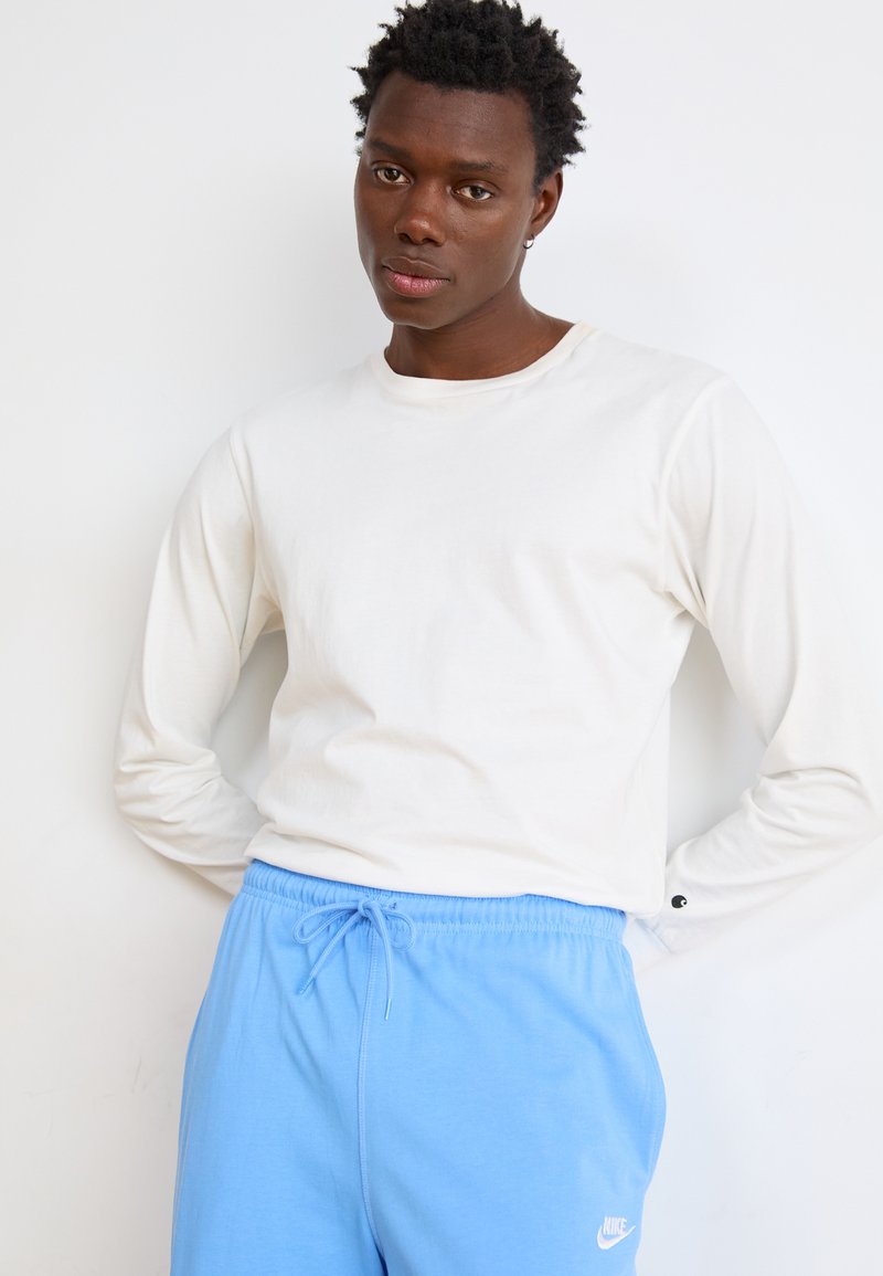 Camisa blanca de manga larga hecha de una tela suave, combinada con pantalones de chándal azul claro con cordón, que cuentan con un pequeño logo blanco en la pierna.