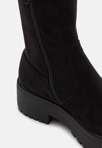 Bota negra de ante hasta la rodilla con suela gruesa de goma, cremallera lateral y textura suave. Presenta un diseño sencillo y detalles mínimos.