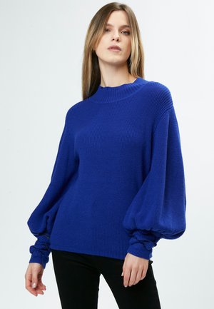 Maglione a coste blu royal con collo alto, maniche a palloncino e polsini arricciati. Abbinato a pantaloni neri, su uno sfondo chiaro.