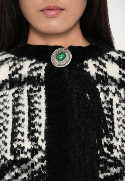 Vêtement à motifs noir et blanc en tissu texturé avec un fermoir rond en argent comportant une pierre verte au centre.