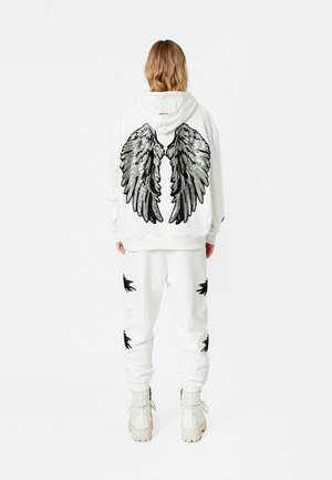 Sudadera blanca con bordado de lentejuelas en forma de alas negras en la parte de atrás, combinada con pantalones de chándal a juego que presentan adornos de alas negras.