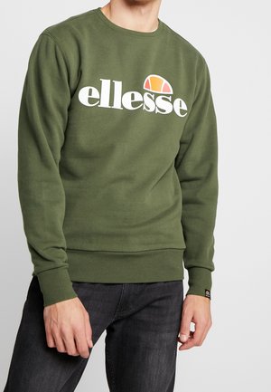 Mężczyzna w oliwkowej bluzie z dużym białym logo „ellesse” i pomarańczowym półokrągłym wzorem, w zestawie z ciemnoszarymi dżinsami.