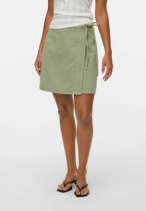 Lichtgroene wikkelrok van zachte stof, met een zijsluiting, een eenvoudige naad en een rechte snit. Draagt zwarte teenslippers.