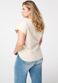 Haut beige à manches courtes avec des accents cloutés aux épaules ; coupe décontractée, fabriqué en tissu doux. Associé à un jean en denim bleu clair.