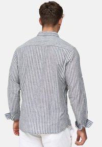 Man die een blauw-wit gestreept overhemd met lange mouwen en een witte broek draagt, van achteren gezien tegen een effen achtergrond.