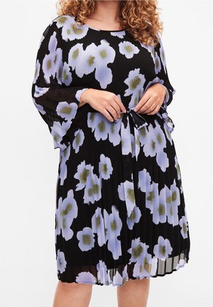 Kobieta ubrana w czarną sukienkę sięgającą kolan, ozdobioną dużymi jasnofioletowymi i zielonymi kwiatowymi wzorami, z długimi rękawami i paskiem wiązanym w talii.