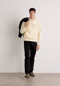 Sudadera amarilla clara con logo en el frente, pantalones negros y zapatos olivo. El modelo sostiene una chaqueta negra sobre un hombro contra un fondo neutro.