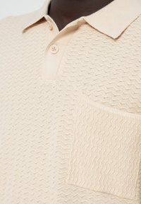 Πλεκτό πόλο μπλουζάκι σε μπεζ χρώμα με υφασμένο σχέδιο, δύο κουμπιά και τσέπη στο στήθος· απαλό ύφασμα με κυματιστό μοτίβο.