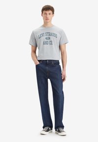 Un jeune homme portant un t-shirt gris Levi Strauss, un jean bleu foncé et des baskets noires se tient avec une main dans la poche sur un fond blanc.