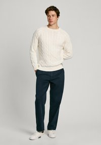 Maglione a treccia crema, pantaloni blu e sneakers bianche. Il maglione presenta un design texture con polsini e orlo a costine.