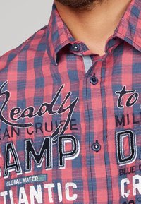 Chemise à carreaux rouge et bleue avec un design à boutons, présentant du texte et des logos brodés. Le tissu semble texturé avec une coupe décontractée.