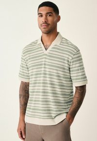 Polo de manga corta a rayas verde claro con cuello tipo v y color crema. Presenta una tela texturizada y un dobladillo acanalado. Tatuajes visibles en el brazo.