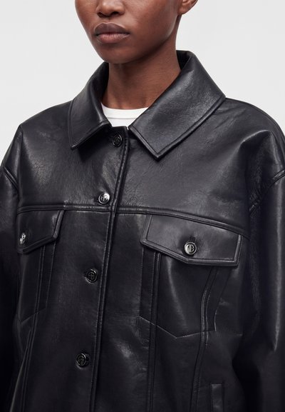 Personne portant une veste en cuir noir brillante avec un large col, des boutons devant et deux poches poitrine, vue des épaules au milieu du torse.