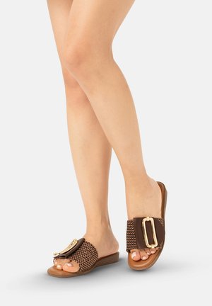 Mujer con sandalias slip-on tejidas marrones con una gran hebilla dorada, de pie con las piernas cruzadas sobre un fondo claro.