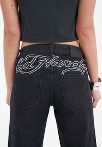 Black denim jeans with back pockets, featuring white embroidered "Ed Hardy" logo on the waistband and a black leather patch above the right pocket.