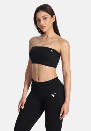 Frau trägt schwarzen trägerlosen Crop-Top und hoch taillierte Leggings mit weißen Logos, posiert vor einem einfachen weißen Hintergrund.