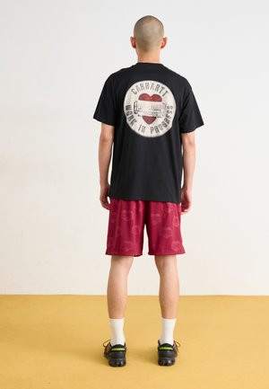 Sort bomuld T-shirt med et rundt motiv på ryggen, kombineret med vinrøde shorts med et diskret mønster, båret med hvide sokker og sorte sko.