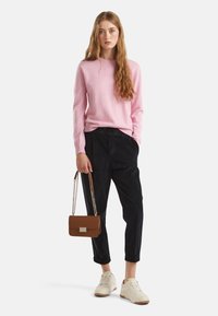 Maglione rosa a maglia, pantaloni in corduroy neri, sneaker beige e una borsa marrone con tracolla a catena. Vista a figura intera con postura eretta.