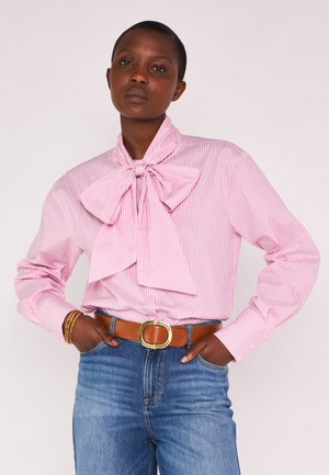 Femme portant un chemisier rayé rose et blanc avec un grand nœud, un jean bleu, une ceinture marron avec une boucle dorée, et des bracelets, debout les mains dans les poches.