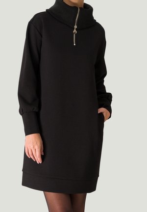 Frau trägt ein schwarzes Langarmkleid mit hohem, geripptem Kragen und halbem Reißverschluss, eine Hand in der Tasche, vor einfarbigem Hintergrund.