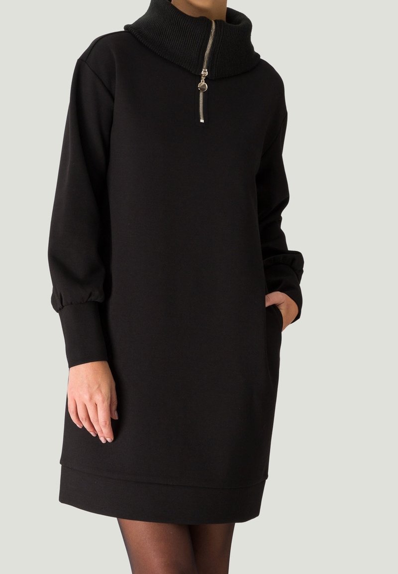 Femme portant une robe noire à manches longues avec un col côtelé haut et une demi-fermeture éclair, une main dans la poche, sur un fond uni.