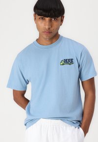 Light blue cotton t-shirt with short sleeves, featuring a graphic logo of a green truck and "HUF TOOL INC" in black lettering on the chest.