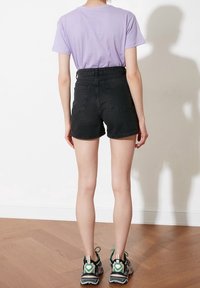 Pantalones cortos de mezclilla negra con cintura alta y bolsillos traseros, combinados con una camiseta lavanda. Persona vistiendo zapatos deportivos grises y verdes.