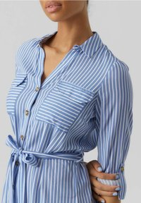 Modro-bílé pruhované šaty košilového střihu s předním zapínáním na knoflíky, límečkem, dvěma náprsními kapsami a zavazovacími pásky v pase. Dlouhé rukávy s knoflíkovými manžetami.