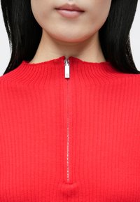 Piros bordázott kötött felső magas nyakrésszel és rövid cipzárral a nyaknál, sima textúrával és látható varrási részletekkel.