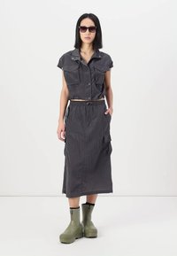 Grijze cropped shirt met twee borstzakken, gecombineerd met een lange grijze rok met zijzakken. Het model draagt groene enkellaarzen en een zonnebril.