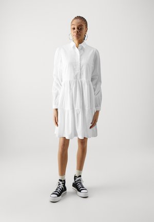 White tiered dress with a button-up front and long sleeves, made from textured fabric. Paired with black high-top sneakers.