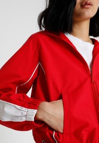 Czerwona kurtka przeciwwietrzna z białymi akcentami, zamek błyskawiczny z przodu, elastyczne mankiety i boczne kieszenie. Gładka faktura i sportowy design.