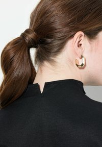Gouden geometrische oorbellen, met een gestructureerd oppervlak, tegen een zwarte top met hoge kraag en een rugventiel detail, die een strak ontwerp toont.