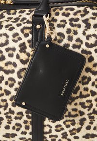 Torba od tkanine s leopard uzorkom s teksturiranim crnim kožnim oznakom. Zlato hardverske akcente dopunjuju dizajn. Oznaka prikazuje naziv brenda.