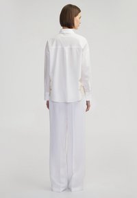 Camisa blanca de manga larga con yugo en la parte posterior y detalles de bordado beige; combinada con pantalones blancos holgados, ambos de tela suave.