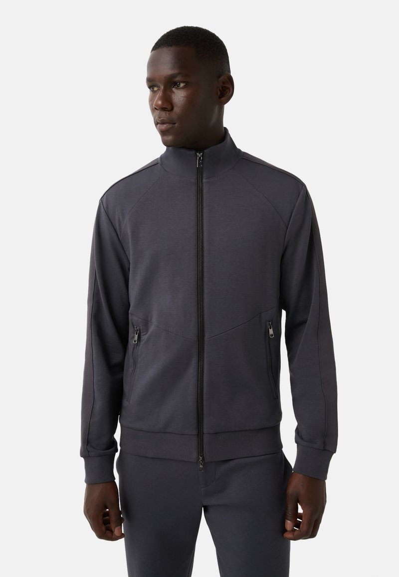 Czarna szara kurtka dresowa z wysokim kołnierzem, pełnym zamkiem błyskawicznym, bocznymi kieszeniami i fakturowanym materiałem. Posiada dopasowane mankiety oraz wyraźne szwy na ramionach.