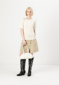 Top bianco a maniche corte a coste, gonna plissettata beige con spacco frontale, stivali neri al ginocchio con ricami floreali bianchi, borsa bianca.