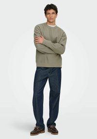 Maglione a maglia testurizzata di un verde oliva chiaro con scollo rotondo, abbinato a jeans blu scuro e scarpe di pelle marroni. Vestibilità dritta.