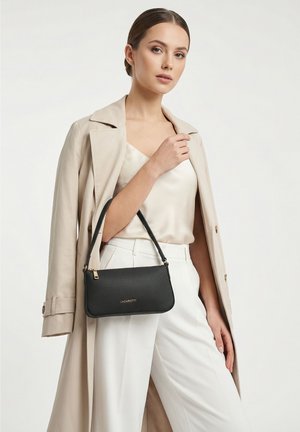 Jonge vrouw draagt een beige jas en een witte satijnen top gecombineerd met een witte broek en houdt een zwarte Lazarotti handtas onder haar arm.