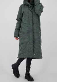 Grön camouflaged puffermantel, knälång med huva, två framdragkedjor och elastiska ärmslut. Bärs över svarta jeans och kraftiga svarta stövlar.