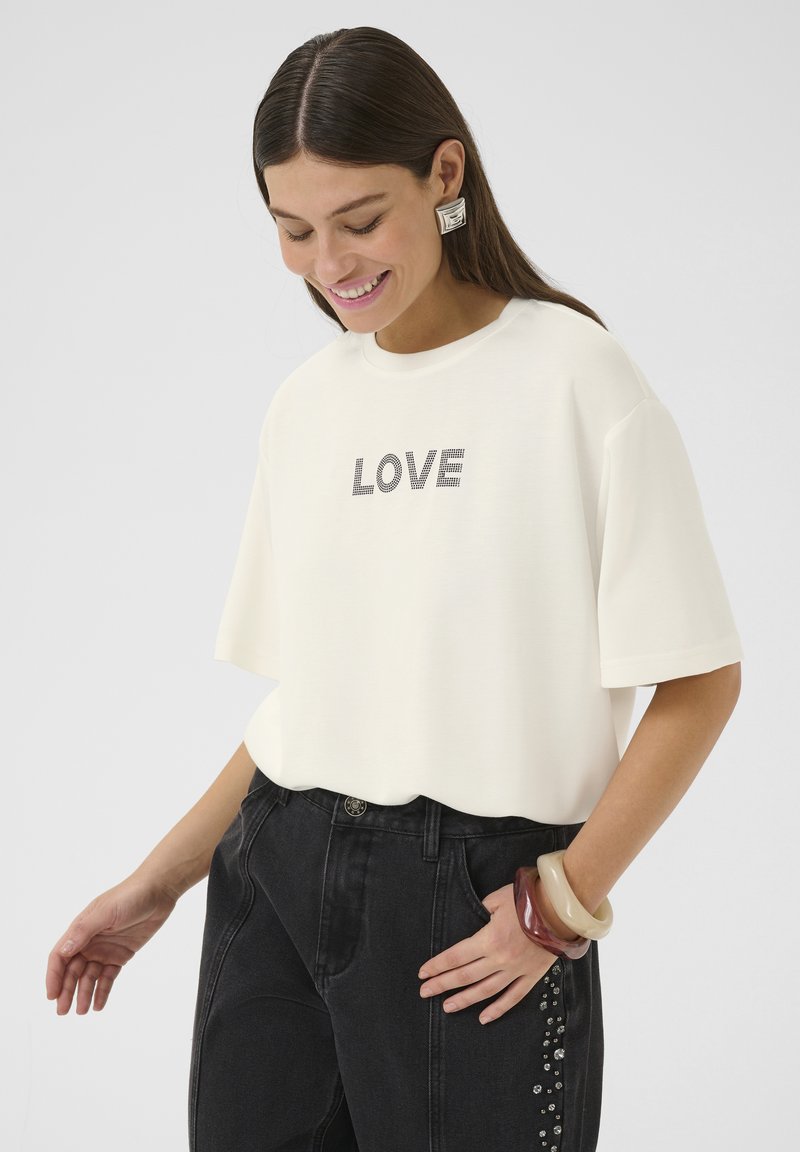 Majica kratkih rukava krem boje s natpisom "LOVE" u crnim sjajnim kamenčićima, u kombinaciji s crnim trapericama s ukrasnim čavlima i masivnim narukvicama.