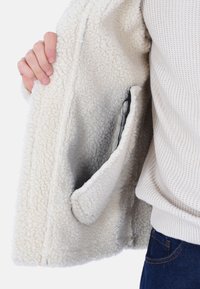 Interno di una giacca in fleece color crema con una texture morbida, caratterizzata da una tasca laterale e un design rilassato. Mano visibile che tiene il bordo.