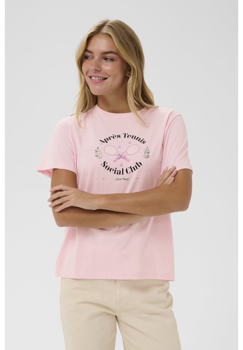 Jauna sieviete ar sakrustotām rokām, ģērbta rozā T-kreklā ar tenisa raketes attēlu un uzrakstu "Après Tennis The Social Club."