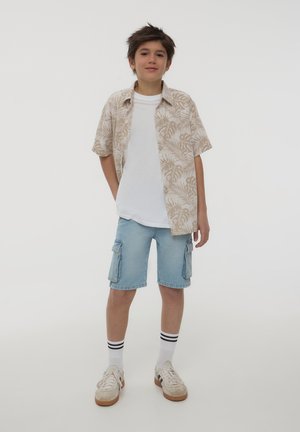 Camicia beige a maniche corte con motivo a foglie sopra una T-shirt bianca, abbinata a pantaloni cargo azzurri chiari e sneaker bianche con suola marrone.